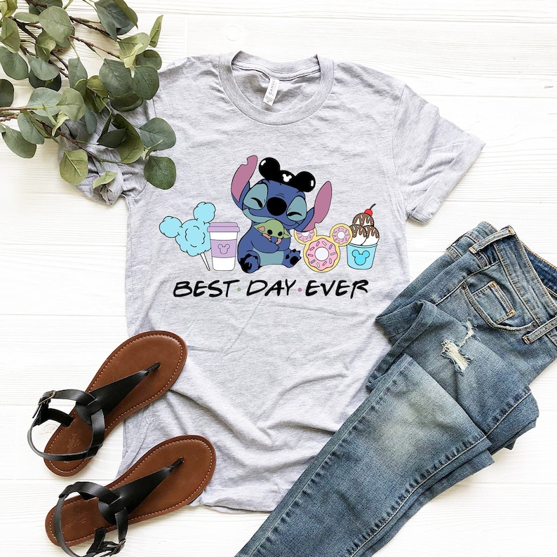 K&ouml;nnte beinhalten: Graues T-Shirt mit einer blauen Stitch-Figur, die eine gr&uuml;ne Baby Yoda-Figur h&auml;lt. Stitch tr&auml;gt Mickey Mouse-Ohren. Das Shirt hat den Text "BEST DAY EVER" und Bilder von Mickey Mouse-Ohren, Zuckerwatte, einer Kaffeetasse, Donuts und einem Mickey Mouse-Eisbecher.