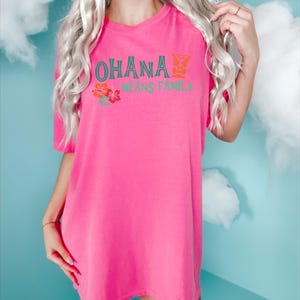 May include: A vibrant pink t-shirt with the words "OHANA MEANS FAMILY" in teal and orange. The shirt features a tropical floral design. The t-shirt is a loose fit and has short sleeves.