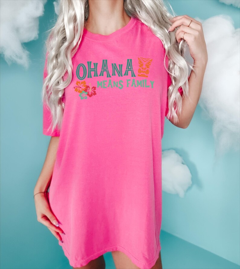 May include: A vibrant pink t-shirt with the words "OHANA MEANS FAMILY" in teal and orange. The shirt features a tropical floral design. The t-shirt is a loose fit and has short sleeves.