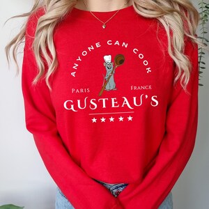 May include: A red long-sleeve sweatshirt with the text "ANYONE CAN COOK" in a circular design. Below the text is an illustration of a rat chef holding a wooden spoon. The word "PARIS" is above "GUSTEAU'S" and "FRANCE" is below it.