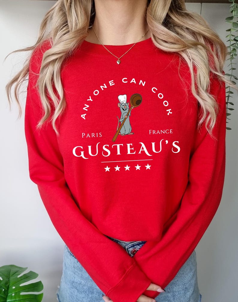May include: A red long-sleeve sweatshirt with the text "ANYONE CAN COOK" in a circular design. Below the text is an illustration of a rat chef holding a wooden spoon. The word "PARIS" is above "GUSTEAU'S" and "FRANCE" is below it.