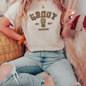 May include: A woman wearing a light beige t-shirt with a brown cartoon character graphic and the text "GROOT EST. 2014 GUARDIANS". She is wearing blue jeans with rips in the knees.