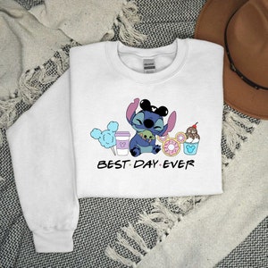 K&ouml;nnte beinhalten: Wei&szlig;er Pullover mit einer blauen Stitch-Figur, die eine gr&uuml;ne Alien-Figur h&auml;lt. Stitch tr&auml;gt Mickey-Mouse-Ohren. Der Text "BEST DAY EVER" ist unter den Figuren gedruckt. Um die Figuren herum befinden sich verschiedene Disney-Motive, darunter ein Mickey-Mouse-Eisbecher, eine Mickey-Mouse-Kaffeetasse, ein Donut und Zuckerwatte.