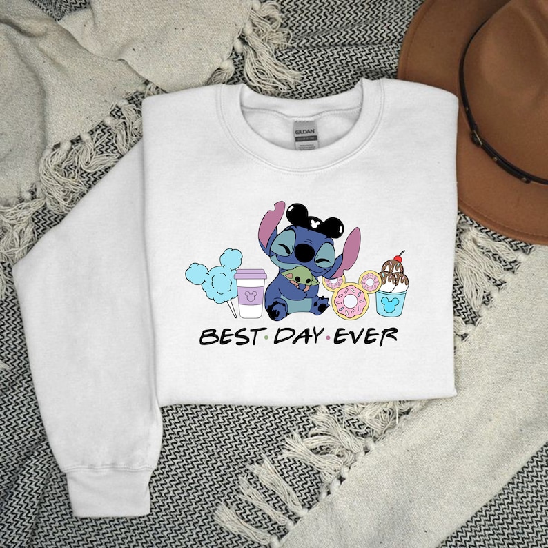 K&ouml;nnte beinhalten: Wei&szlig;er Pullover mit einer blauen Stitch-Figur, die eine gr&uuml;ne Alien-Figur h&auml;lt. Stitch tr&auml;gt Mickey-Mouse-Ohren. Der Text "BEST DAY EVER" ist unter den Figuren gedruckt. Um die Figuren herum befinden sich verschiedene Disney-Motive, darunter ein Mickey-Mouse-Eisbecher, eine Mickey-Mouse-Kaffeetasse, ein Donut und Zuckerwatte.