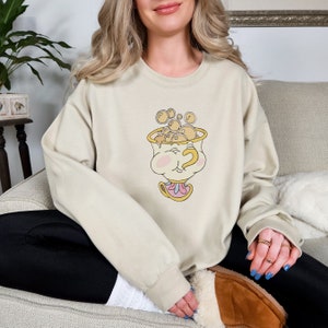 Puede incluir: Una sudadera beige con una imagen de dibujos animados de una taza de t&eacute; con una cara y un patr&oacute;n de burbujas amarillas en espiral. La taza de t&eacute; lleva un sombrero rosa y amarillo.