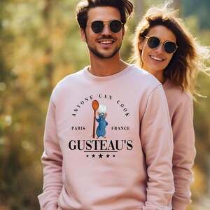 May include: A light pink sweatshirt with the text "ANYONE CAN COOK" above a cartoon rat chef holding a spoon. Below the rat is the text "GUSTEAU'S" with stars. The sweatshirt is worn by a person wearing sunglasses.