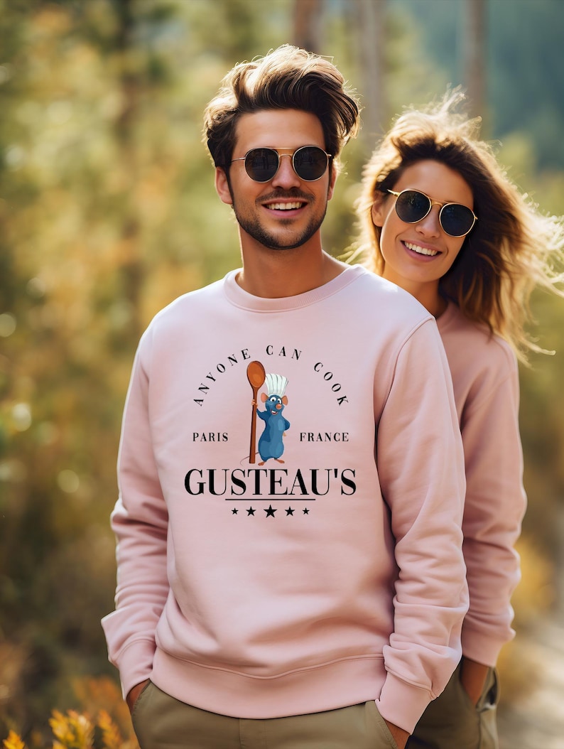 May include: A light pink sweatshirt with the text "ANYONE CAN COOK" above a cartoon rat chef holding a spoon. Below the rat is the text "GUSTEAU'S" with stars. The sweatshirt is worn by a person wearing sunglasses.
