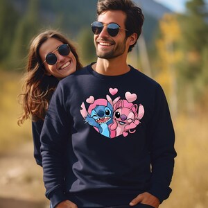 Puede incluir: Sudadera azul marino con una imagen de dibujos animados de Stitch y Angel, rodeados de corazones rosas. El dise&ntilde;o est&aacute; centrado en el pecho. La sudadera la lleva una persona con gafas de sol.