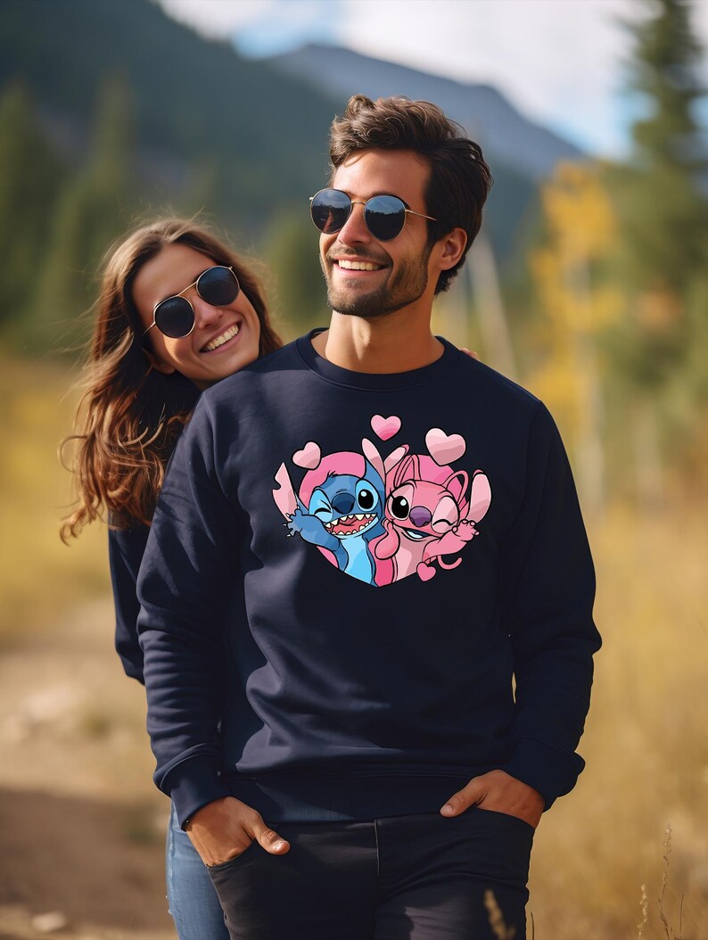 Puede incluir: Sudadera azul marino con una imagen de dibujos animados de Stitch y Angel, rodeados de corazones rosas. El dise&ntilde;o est&aacute; centrado en el pecho. La sudadera la lleva una persona con gafas de sol.