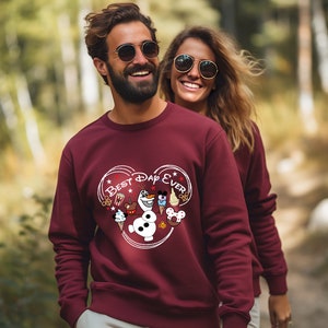 May include: A maroon sweatshirt with a white heart design featuring the text "Best Day Ever" and various Disney characters and icons, including Mickey Mouse, Olaf, and a popcorn bucket.