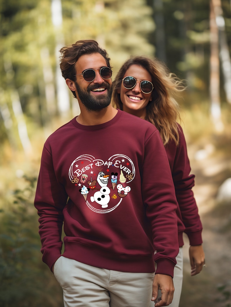 May include: A maroon sweatshirt with a white heart design featuring the text "Best Day Ever" and various Disney characters and icons, including Mickey Mouse, Olaf, and a popcorn bucket.