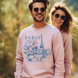 Op de afbeelding: Een roze sweatshirt met een blauwe afbeelding van Mickey Mouse en Minnie Mouse voor de Eiffeltoren. Het sweatshirt heeft het woord "PARIS" op de voorkant.
