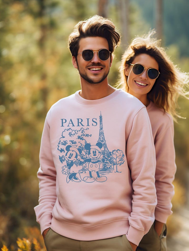 Op de afbeelding: Een roze sweatshirt met een blauwe afbeelding van Mickey Mouse en Minnie Mouse voor de Eiffeltoren. Het sweatshirt heeft het woord "PARIS" op de voorkant.