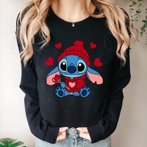 May include: Black crewneck sweatshirt featuring a cartoon character wearing a red knit hat and sweater, holding a heart-shaped lollipop. Red hearts are scattered around the character. The character is blue with pink ears and feet.