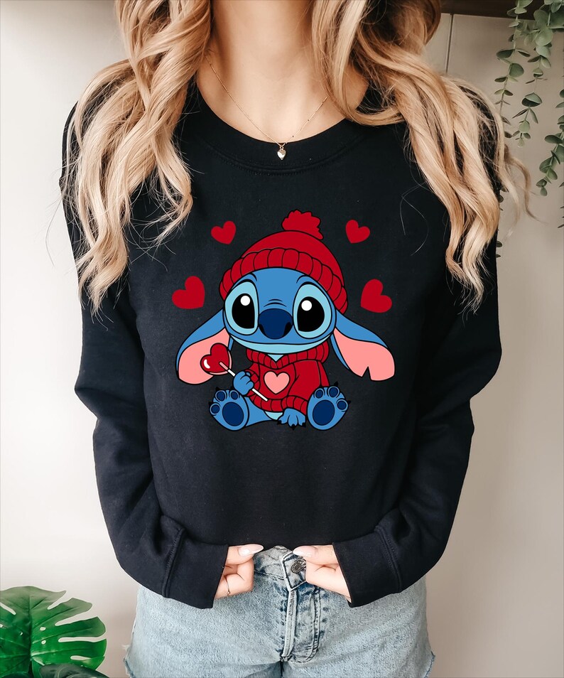 May include: Black crewneck sweatshirt featuring a cartoon character wearing a red knit hat and sweater, holding a heart-shaped lollipop. Red hearts are scattered around the character. The character is blue with pink ears and feet.