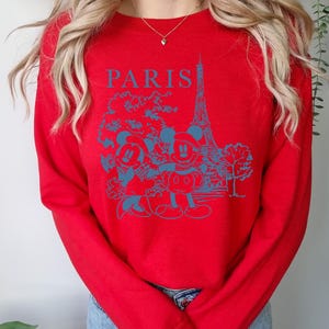 Op de afbeelding: Rode longsleeve trui met een blauwe grafische print van Mickey Mouse en Minnie Mouse voor de Eiffeltoren. De tekst "PARIS" is boven de print gedrukt.