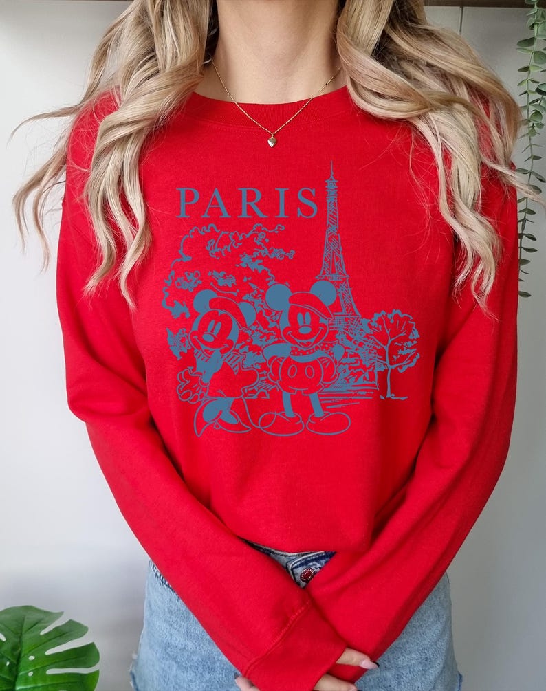 Op de afbeelding: Rode longsleeve trui met een blauwe grafische print van Mickey Mouse en Minnie Mouse voor de Eiffeltoren. De tekst "PARIS" is boven de print gedrukt.