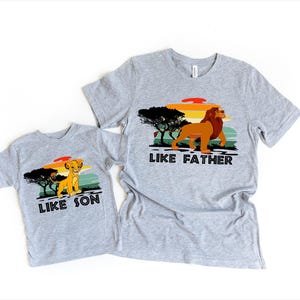 May include: Two grey t-shirts with a retro sunset graphic and lion images. The larger shirt says "Like Father" and the smaller shirt says "Like Son".