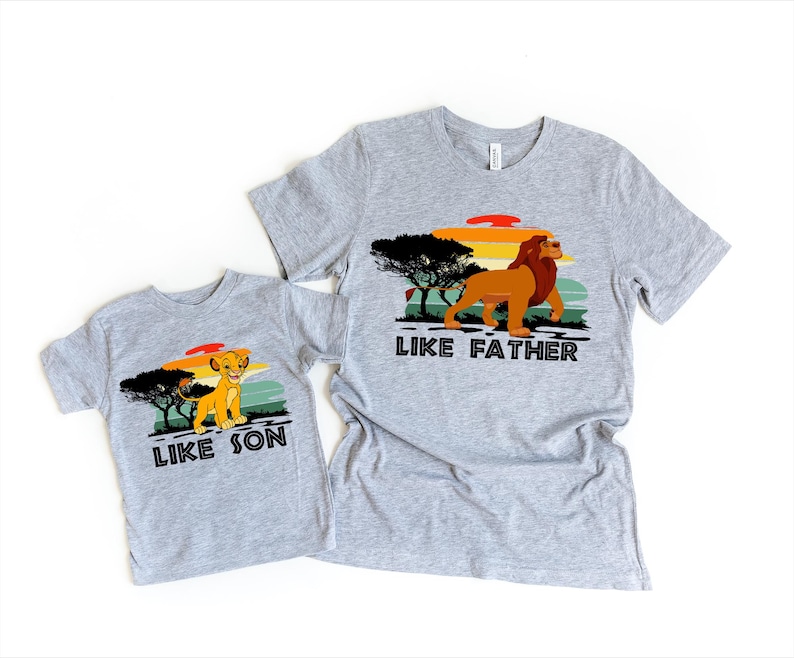 May include: Two grey t-shirts with a retro sunset graphic and lion images. The larger shirt says "Like Father" and the smaller shirt says "Like Son".