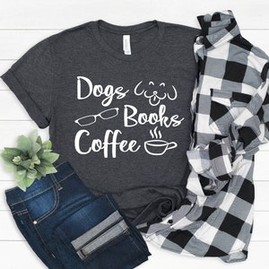 May include: A dark charcoal gray t-shirt featuring white text that reads Dogs, Books, and Coffee, accompanied by white line art icons of a dog face, eyeglasses, and a steaming coffee cup, paired with dark blue jeans and a black and white plaid shirt.
