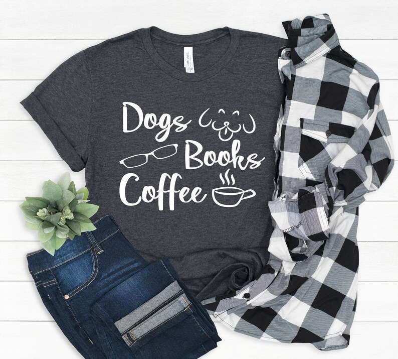 May include: A dark charcoal gray t-shirt featuring white text that reads Dogs, Books, and Coffee, accompanied by white line art icons of a dog face, eyeglasses, and a steaming coffee cup, paired with dark blue jeans and a black and white plaid shirt.