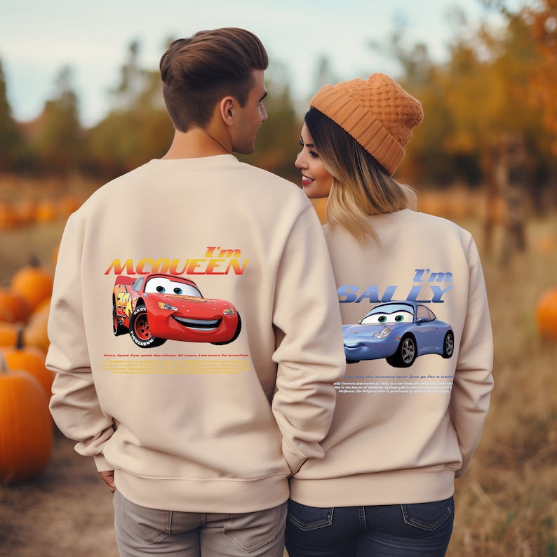 May include: A couple wearing matching beige sweatshirts with a Cars theme. The man's sweatshirt features the text "I'm McQueen" and a red race car. The woman's sweatshirt features the text "I'm Sally" and a blue race car.