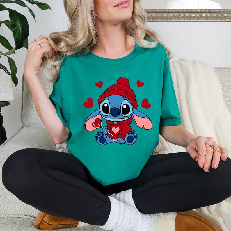 May include: Teal green t-shirt featuring a cartoon character wearing a red hat and sweater, surrounded by red hearts. The character is holding a heart-shaped lollipop. The shirt is a casual, comfortable style.