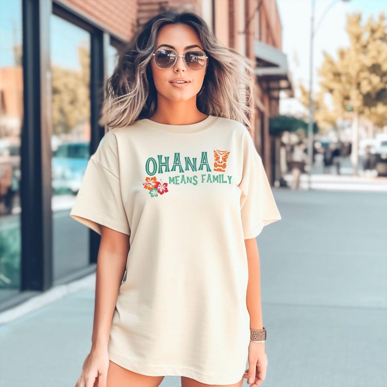 May include: A cream-coloured t-shirt with the text "OHANA MEANS FAMILY" in green and red. The shirt features a floral design and a stylized graphic. The t-shirt is made of a soft material.
