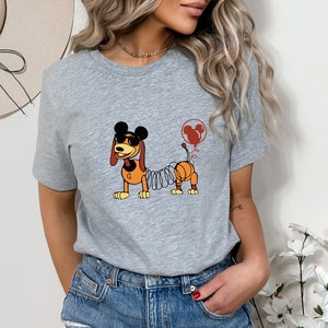 May include: A gray t-shirt with a cartoon image of a dog with a spring body and Mickey Mouse ears. The dog is wearing sunglasses and has a red balloon with Mickey Mouse ears attached to its tail.