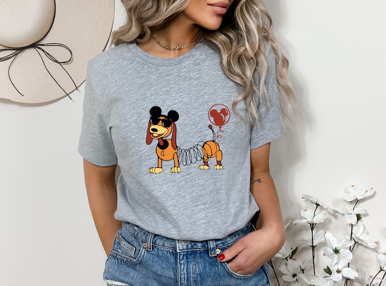 May include: A gray t-shirt with a cartoon image of a dog with a spring body and Mickey Mouse ears. The dog is wearing sunglasses and has a red balloon with Mickey Mouse ears attached to its tail.