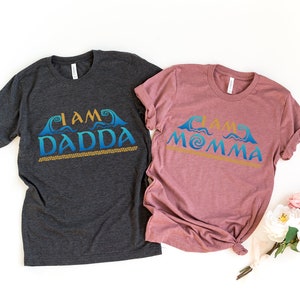 May include: Two t-shirts with a Moana-inspired design. The dark grey shirt says "I am Dadda" and the mauve shirt says "I am Momma". Both shirts feature a blue and gold wave design.