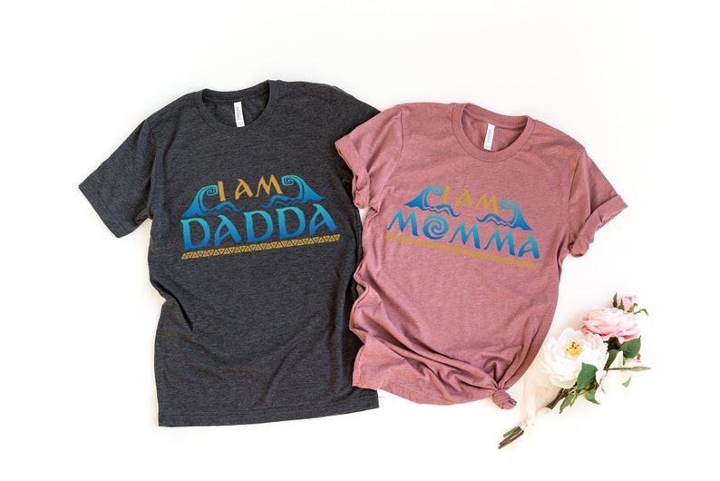 May include: Two t-shirts with a Moana-inspired design. The dark grey shirt says "I am Dadda" and the mauve shirt says "I am Momma". Both shirts feature a blue and gold wave design.