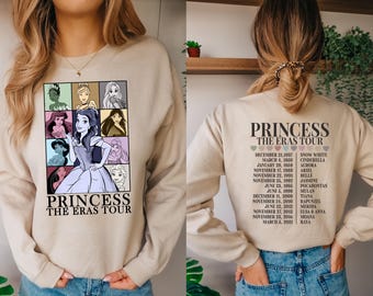 Może przedstawiać: Beżowa bluza z nadrukiem graficznym przedstawiającym księżniczki Disneya i tekstem "Princess The Eras Tour". Na plecach bluzy znajduje się lista księżniczek Disneya i odpowiadające im daty trasy koncertowej.