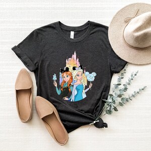 Op de afbeelding: Een donkergrijs T-shirt met een afbeelding van de Disney-prinsessen Anna en Elsa voor het kasteel van Assepoester. Ze houden Mickey Mouse-oren en lekkernijen vast.