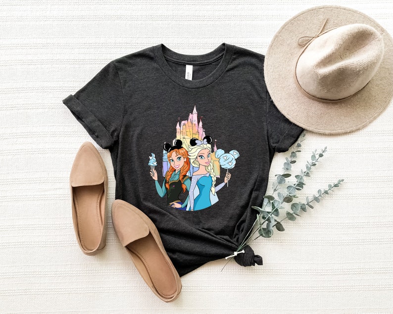 Op de afbeelding: Een donkergrijs T-shirt met een afbeelding van de Disney-prinsessen Anna en Elsa voor het kasteel van Assepoester. Ze houden Mickey Mouse-oren en lekkernijen vast.