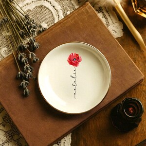 May include: A small, round, off-white ceramic plate with a gold rim. The plate features a watercolor-style red poppy and the handwritten word "netelie". The plate rests on a brown book.