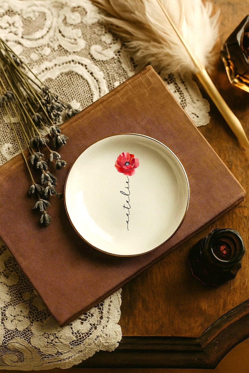 May include: A small, round, off-white ceramic plate with a gold rim. The plate features a watercolor-style red poppy and the handwritten word "netelie". The plate rests on a brown book.