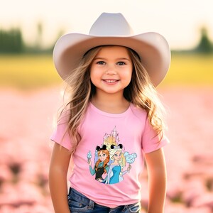 Op de afbeelding: Een jong meisje draagt een roze T-shirt met een afbeelding van het Disney-kasteel, Anna en Elsa uit Frozen, en Mickey Mouse-oren. Ze draagt ook een witte cowboyhoed en een blauwe jeans.