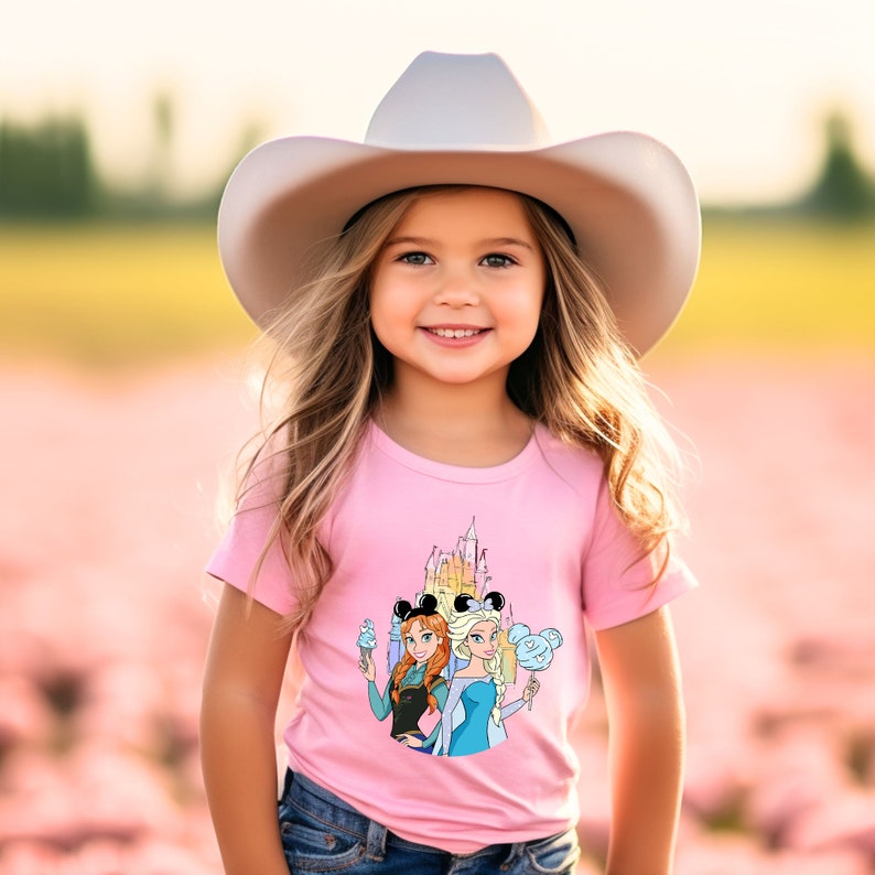 Op de afbeelding: Een jong meisje draagt een roze T-shirt met een afbeelding van het Disney-kasteel, Anna en Elsa uit Frozen, en Mickey Mouse-oren. Ze draagt ook een witte cowboyhoed en een blauwe jeans.