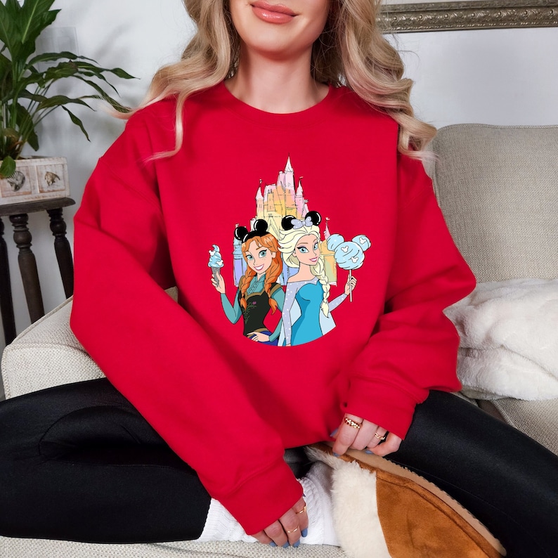 Op de afbeelding: Rood sweatshirt met een grafische print van het Disney-kasteel, Elsa en Anna van Frozen, die Mickey Mouse-oren dragen en ijs en suikerspin houden.