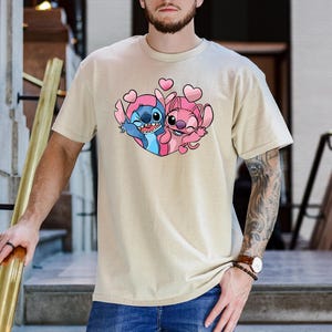 Puede incluir: Camiseta beige con un gr&aacute;fico colorido de Stitch y Angel, rodeados de corazones rosas. Los personajes de dibujos animados se abrazan, Stitch en azul y rosa, y Angel en rosa. La camiseta tiene mangas cortas y cuello redondo.