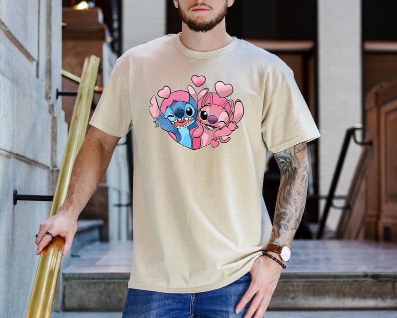 Puede incluir: Camiseta beige con un gr&aacute;fico colorido de Stitch y Angel, rodeados de corazones rosas. Los personajes de dibujos animados se abrazan, Stitch en azul y rosa, y Angel en rosa. La camiseta tiene mangas cortas y cuello redondo.