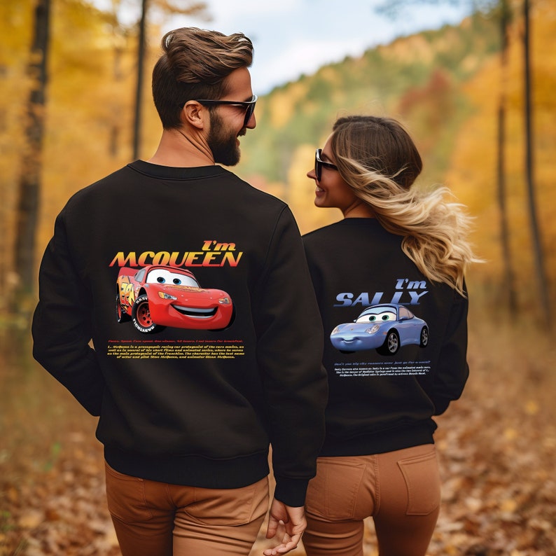 May include: A couple wearing black sweatshirts with a graphic print of the characters Lightning McQueen and Sally from the Disney Pixar movie Cars. The man's sweatshirt says "I'm McQueen" and the woman's sweatshirt says "I'm Sally".
