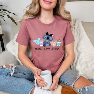 K&ouml;nnte beinhalten: Ein mauvefarbenes T-Shirt mit einem Cartoon-Stitch, der Mickey-Mouse-Ohren tr&auml;gt und ein gr&uuml;nes Alien h&auml;lt. Das Shirt tr&auml;gt den Aufdruck "Best Day Ever".
