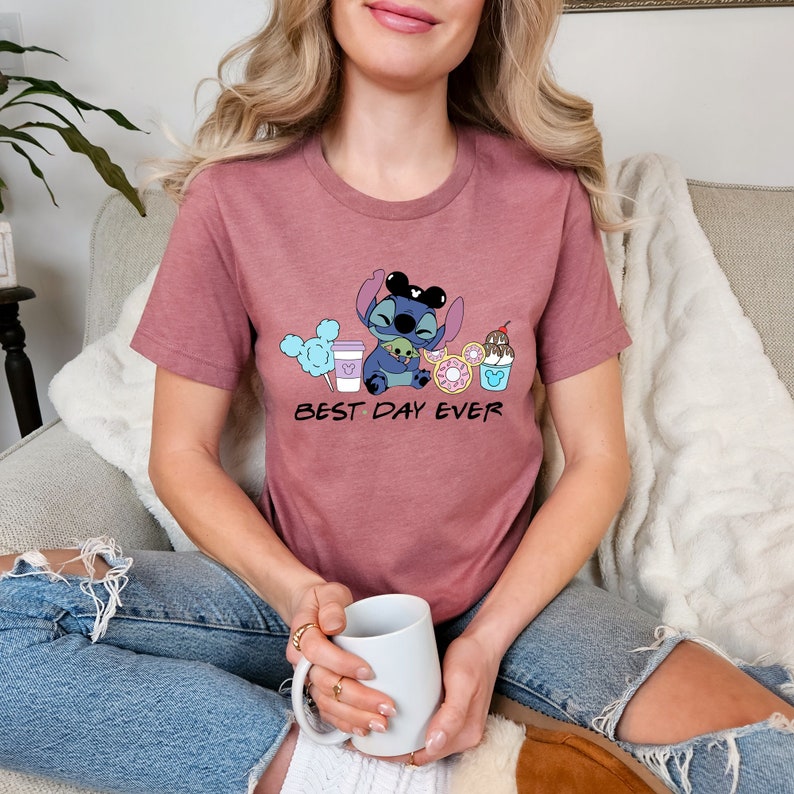 K&ouml;nnte beinhalten: Ein mauvefarbenes T-Shirt mit einem Cartoon-Stitch, der Mickey-Mouse-Ohren tr&auml;gt und ein gr&uuml;nes Alien h&auml;lt. Das Shirt tr&auml;gt den Aufdruck "Best Day Ever".