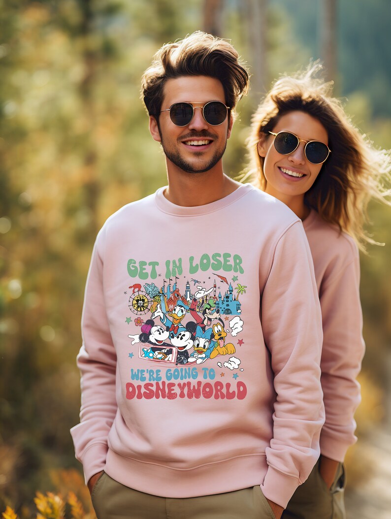 Puede incluir: Una sudadera rosa con un estampado gr&aacute;fico de Mickey Mouse y otros personajes de Disney. El texto en la sudadera dice "Get in loser, we're going to Disney World."