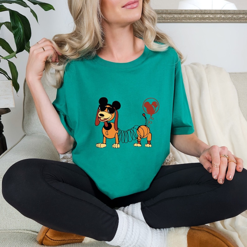 May include: A teal green t-shirt with a cartoon image of a dog with a spring for a tail. The dog is wearing sunglasses and a Mickey Mouse hat. The dog is holding a red balloon with a Mickey Mouse head on it.