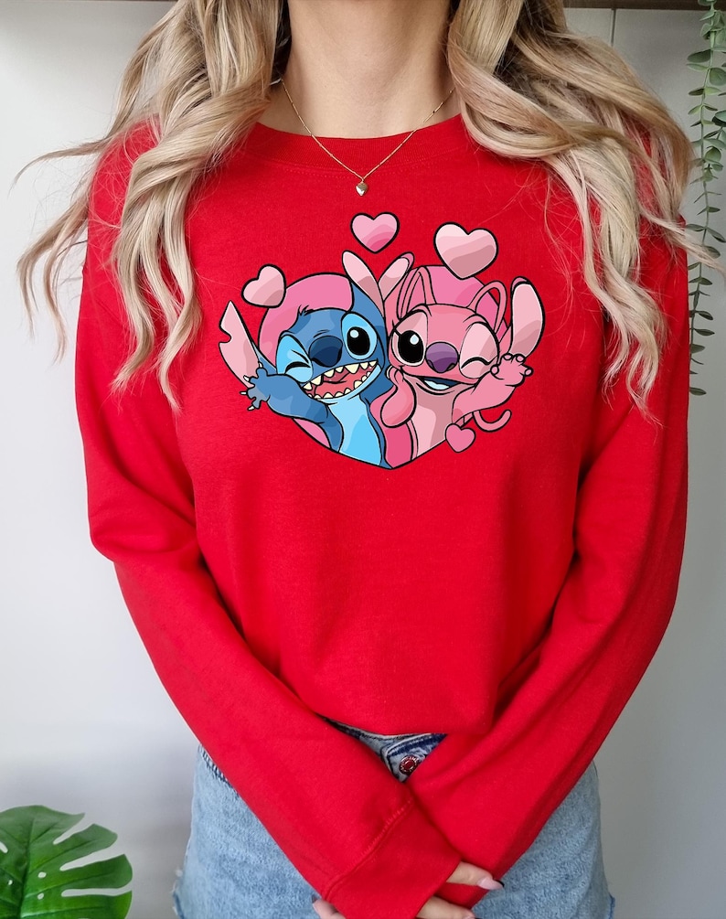 Puede incluir: Una sudadera roja con un gr&aacute;fico de los personajes Stitch y Angel de Lilo & Stitch, rodeados de corazones rosas. Los personajes de dibujos animados se abrazan, Stitch gui&ntilde;a un ojo. La sudadera es de color rojo liso.