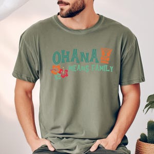 May include: Olive green t-shirt with the words "OHANA MEANS FAMILY" in teal and orange. The shirt features a floral design with hibiscus flowers. The t-shirt is made of a soft, comfortable material.