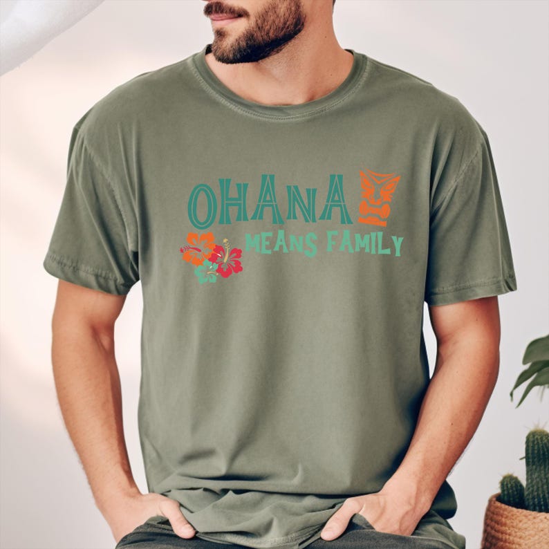May include: Olive green t-shirt with the words "OHANA MEANS FAMILY" in teal and orange. The shirt features a floral design with hibiscus flowers. The t-shirt is made of a soft, comfortable material.